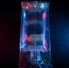 The image shows a medical intravenous (IV) fluid bag photographed under dramatic blue and red/pink lighting against a dark background. The transparent plastic bag contains a clear liquid and features standard IV administration ports at the bottom. The artistic lighting gives the medical equipment a cinematic, stylized appearance.