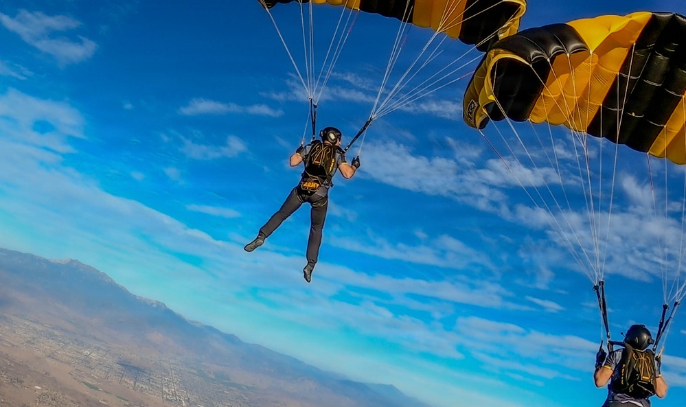 Two skydivers are descending under deployed black and gold parachutes against a bright blue sky with scattered clouds. The left skydiver is wearing gear marked with 'ARMY' in yellow lettering, indicating affiliation with a U.S. Army parachute team, likely the Golden Knights. A mountainous landscape and urban area are visible far below.