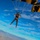 Golden Knights Defend Canopy Formation Title at Perris Two skydivers are descending under deployed black and gold parachutes against a bright blue sky with scattered clouds. The left skydiver is wearing gear marked with 'ARMY' in yellow lettering, indicating affiliation with a U.S. Army parachute team, likely the Golden Knights. A mountainous landscape and urban area are visible far below.