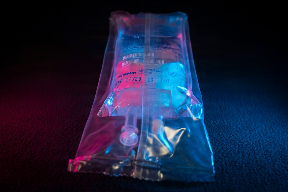 The image shows a sealed medical IV bag or infusion pouch, manufactured by B. Braun, photographed under dramatic pink and blue lighting against a dark background. The transparent outer packaging contains an inner IV bag with visible tubing and connectors. The expiration date visible on the label reads 12/23, and a LOT number is partially visible.