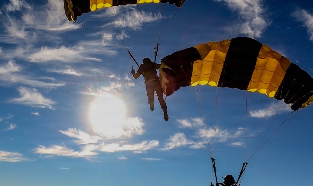 The image captures multiple skydivers descending under deployed parachutes against a bright blue sky with scattered clouds. The parachutes are distinctively colored in yellow and black stripes, suggesting a coordinated team or demonstration jump. The photo is taken from a low angle looking upward, with the sun creating a dramatic backlit silhouette effect.