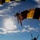 Golden Knights Defend Canopy Formation Title at Perris The image captures multiple skydivers descending under deployed parachutes against a bright blue sky with scattered clouds. The parachutes are distinctively colored in yellow and black stripes, suggesting a coordinated team or demonstration jump. The photo is taken from a low angle looking upward, with the sun creating a dramatic backlit silhouette effect.