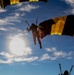 The image captures multiple skydivers descending under deployed parachutes against a bright blue sky with scattered clouds. The parachutes are distinctively colored in yellow and black stripes, suggesting a coordinated team or demonstration jump. The photo is taken from a low angle looking upward, with the sun creating a dramatic backlit silhouette effect.