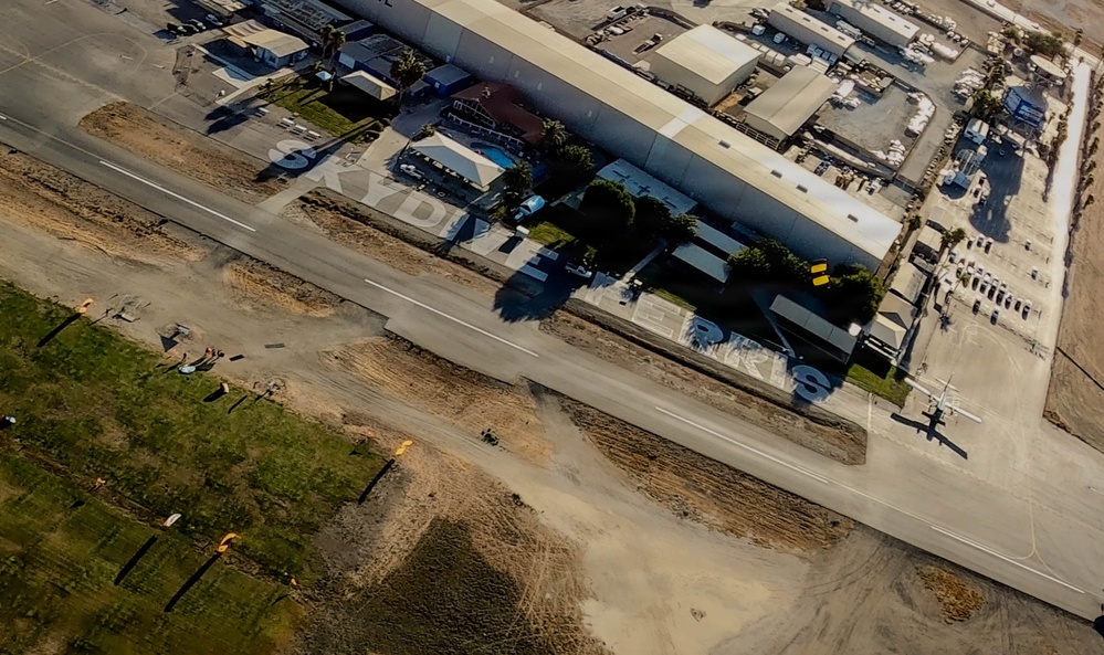 An aerial photograph of a small general aviation airport facility, likely a skydiving operation based on visible text reading 'SKYDIVE' on the ground/buildings. The image shows a runway or taxiway, hangars, support buildings, and at least one small aircraft visible on the tarmac. The surrounding area includes sandy/disturbed terrain, green grass patches, and a residential or commercial zone adjacent to the airfield.