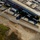 Golden Knights Defend Canopy Formation Title at Perris An aerial photograph of a small general aviation airport facility, likely a skydiving operation based on visible text reading 'SKYDIVE' on the ground/buildings. The image shows a runway or taxiway, hangars, support buildings, and at least one small aircraft visible on the tarmac. The surrounding area includes sandy/disturbed terrain, green grass patches, and a residential or commercial zone adjacent to the airfield.