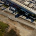 An aerial photograph of a small general aviation airport facility, likely a skydiving operation based on visible text reading 'SKYDIVE' on the ground/buildings. The image shows a runway or taxiway, hangars, support buildings, and at least one small aircraft visible on the tarmac. The surrounding area includes sandy/disturbed terrain, green grass patches, and a residential or commercial zone adjacent to the airfield.