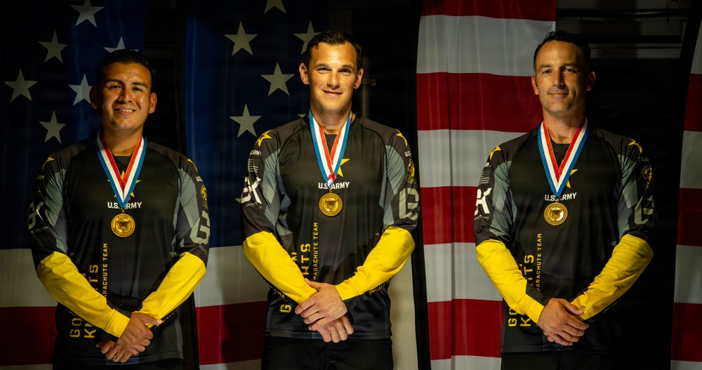 Three individuals wearing U.S. Army Golden Knights Parachute Team uniforms stand in front of an American flag backdrop. Each person is wearing a gold medal on a red, white, and blue ribbon around their neck. The image appears to be an official award or recognition photograph.
