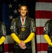 Three individuals wearing U.S. Army Golden Knights Parachute Team uniforms stand in front of an American flag backdrop. Each person is wearing a gold medal on a red, white, and blue ribbon around their neck. The image appears to be an official award or recognition photograph.