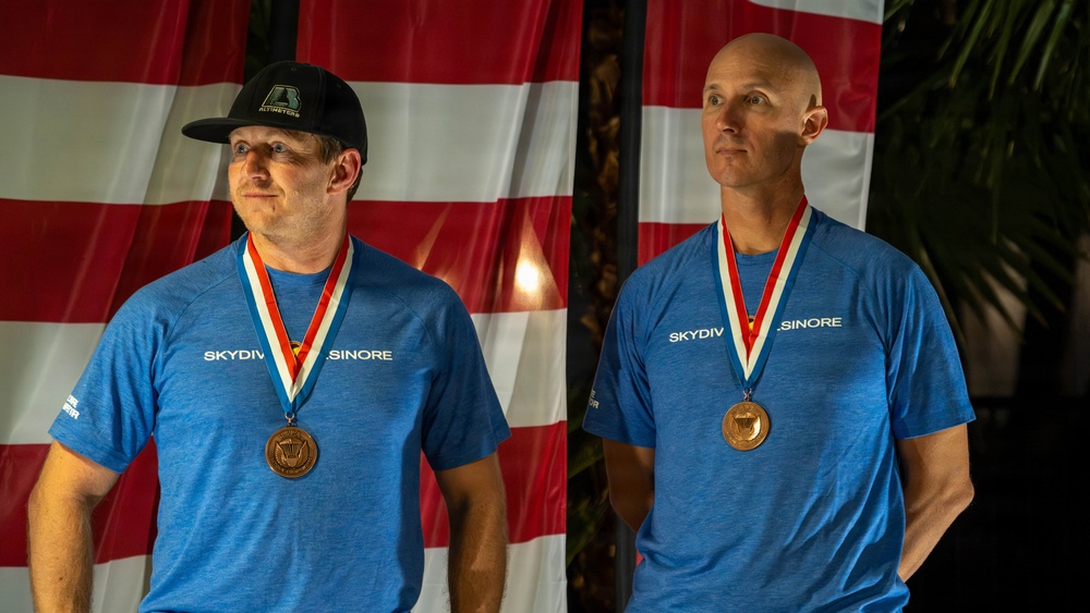 Two men are photographed side by side, each wearing matching blue 'Skydive Elsinore' t-shirts and bronze medals on red, white, and blue ribbons. They are posed in front of what appears to be an American flag backdrop. The image appears to be a composite or split photo taken at an awards ceremony or competition.