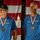 Golden Knights Defend Canopy Formation Title at Perris Two men are photographed side by side, each wearing matching blue 'Skydive Elsinore' t-shirts and bronze medals on red, white, and blue ribbons. They are posed in front of what appears to be an American flag backdrop. The image appears to be a composite or split photo taken at an awards ceremony or competition.