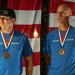Two men are photographed side by side, each wearing matching blue 'Skydive Elsinore' t-shirts and bronze medals on red, white, and blue ribbons. They are posed in front of what appears to be an American flag backdrop. The image appears to be a composite or split photo taken at an awards ceremony or competition.