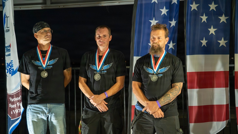Three adult males stand side by side wearing matching black 'Leading Edge Technology' t-shirts and medals with red, white, and blue ribbons, suggesting a competition or award ceremony. They are positioned in front of an American flag and a sponsor banner, indicating a formal recognition event. All three men wear purple wristbands and stand in a composed, attentive posture.