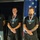 Golden Knights Defend Canopy Formation Title at Perris Three adult males stand side by side wearing matching black 'Leading Edge Technology' t-shirts and medals with red, white, and blue ribbons, suggesting a competition or award ceremony. They are positioned in front of an American flag and a sponsor banner, indicating a formal recognition event. All three men wear purple wristbands and stand in a composed, attentive posture.