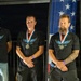 Three adult males stand side by side wearing matching black 'Leading Edge Technology' t-shirts and medals with red, white, and blue ribbons, suggesting a competition or award ceremony. They are positioned in front of an American flag and a sponsor banner, indicating a formal recognition event. All three men wear purple wristbands and stand in a composed, attentive posture.