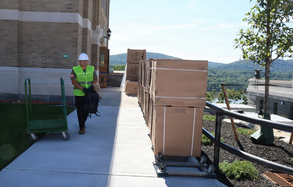 A worker in a high-visibility green vest and white hard hat walks along a sidewalk beside a brick building, passing a row of large cardboard boxes on wheeled carts. The boxes are branded with 'Krug' and appear to be furniture or equipment deliveries. The setting appears to be a construction or renovation site with a scenic river valley visible in the background.