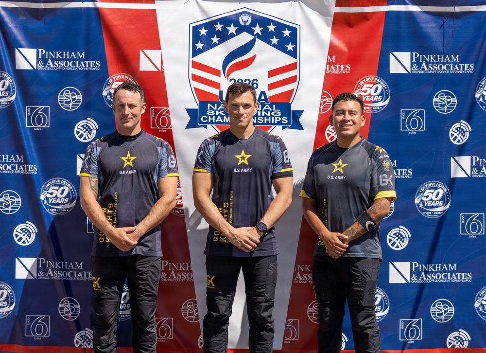 Three individuals wearing matching U.S. Army uniforms pose together in front of a sponsor backdrop at the 2026 USA National Skydiving Championships. Their shirts identify them as members of the Golden Knights Parachute Team. The event is sponsored by Pinkham &amp; Associates (Orange County Divorce Attorneys APLC) and Skydive Perris (celebrating 50 years), among others.