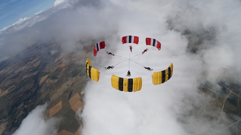 A group of skydivers has formed a circular canopy formation high above a patchwork agricultural landscape. The formation consists of approximately 8-9 parachutists with two distinct canopy color schemes — yellow with black stripes and red with white/black stripes — arranged in a ring pattern. The image is taken from above, likely by a camera flyer, with clouds partially surrounding the formation.