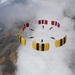 A group of skydivers has formed a circular canopy formation high above a patchwork agricultural landscape. The formation consists of approximately 8-9 parachutists with two distinct canopy color schemes — yellow with black stripes and red with white/black stripes — arranged in a ring pattern. The image is taken from above, likely by a camera flyer, with clouds partially surrounding the formation.