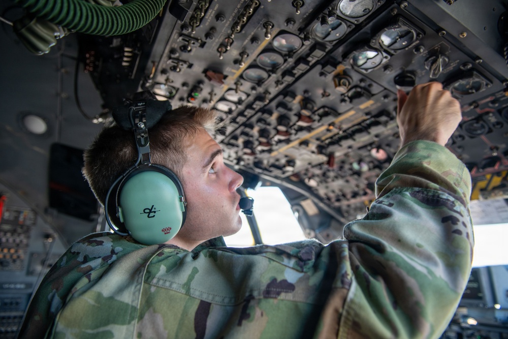 A young military personnel in camouflage uniform is operating or inspecting an overhead control panel inside a military aircraft cockpit. He is wearing mint-green aviation headsets and reaching upward to manipulate switches or controls on the overhead panel. The image is taken from a low angle, emphasizing the dense array of instruments and controls on the overhead console.