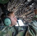 A young military personnel in camouflage uniform is operating or inspecting an overhead control panel inside a military aircraft cockpit. He is wearing mint-green aviation headsets and reaching upward to manipulate switches or controls on the overhead panel. The image is taken from a low angle, emphasizing the dense array of instruments and controls on the overhead console.