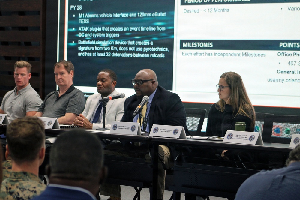 A panel discussion or briefing is taking place at a conference table with five panelists seated in front of a large projected presentation screen. The presentation displays military-related content including FY28 objectives, milestones, and period of performance details. Name placards on the table identify roles including 'Project Manager Training Devices,' 'Project Manager Simulations,' and 'Project Lead Threat Systems Management Office.'