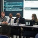A panel discussion or briefing is taking place at a conference table with five panelists seated in front of a large projected presentation screen. The presentation displays military-related content including FY28 objectives, milestones, and period of performance details. Name placards on the table identify roles including 'Project Manager Training Devices,' 'Project Manager Simulations,' and 'Project Lead Threat Systems Management Office.'