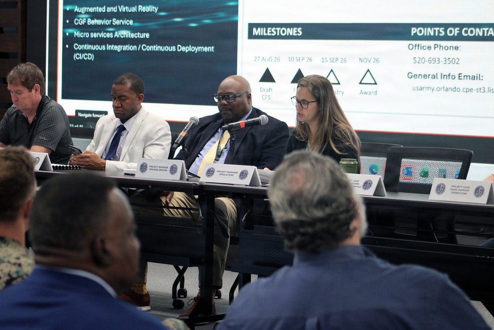 A formal panel discussion or briefing is taking place, likely a government or military acquisition meeting. Four panelists are seated at a long table with name placards, facing an audience, with a presentation slide visible behind them showing milestones and points of contact. The slide references dates in 2026 and topics including Augmented/Virtual Reality, CGF Behavior Service, and CI/CD.