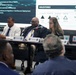 A formal panel discussion or briefing is taking place, likely a government or military acquisition meeting. Four panelists are seated at a long table with name placards, facing an audience, with a presentation slide visible behind them showing milestones and points of contact. The slide references dates in 2026 and topics including Augmented/Virtual Reality, CGF Behavior Service, and CI/CD.