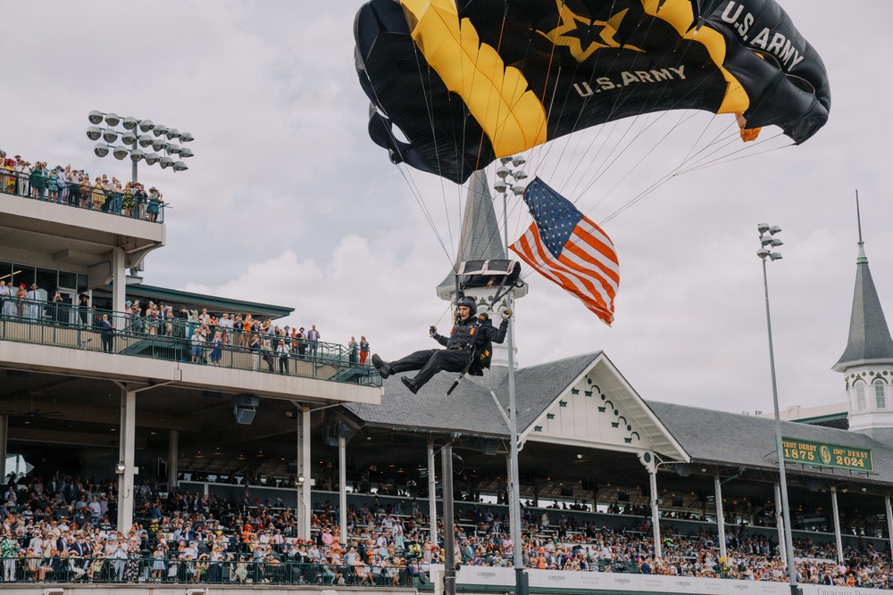 A U.S. Army parachutist is descending into Churchill Downs during what appears to be the 150th Kentucky Derby (2024), as indicated by signage reading 'First Derby 1875' and '150th Derby 2024'. The soldier is carrying an American flag while using a black and gold U.S. Army branded parachute. Large crowds of spectators fill the grandstands watching the ceremonial skydive.