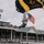 USAPT Golden Knights Parachutist Jump into Kentucky Derby A U.S. Army parachutist is descending into Churchill Downs during what appears to be the 150th Kentucky Derby (2024), as indicated by signage reading 'First Derby 1875' and '150th Derby 2024'. The soldier is carrying an American flag while using a black and gold U.S. Army branded parachute. Large crowds of spectators fill the grandstands watching the ceremonial skydive.