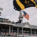 A U.S. Army parachutist is descending into Churchill Downs during what appears to be the 150th Kentucky Derby (2024), as indicated by signage reading 'First Derby 1875' and '150th Derby 2024'. The soldier is carrying an American flag while using a black and gold U.S. Army branded parachute. Large crowds of spectators fill the grandstands watching the ceremonial skydive.