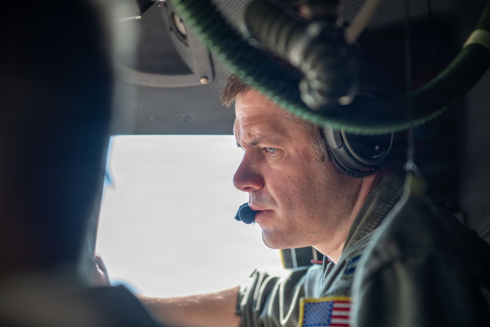 A middle-aged male military aviator is photographed in close profile inside what appears to be a military helicopter cockpit. He is wearing a flight helmet with communications headset and a microphone boom, along with a military flight suit bearing a U.S. flag patch on the shoulder. The image is taken from a tight angle, suggesting the photographer is seated nearby within the aircraft.