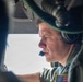A middle-aged male military aviator is photographed in close profile inside what appears to be a military helicopter cockpit. He is wearing a flight helmet with communications headset and a microphone boom, along with a military flight suit bearing a U.S. flag patch on the shoulder. The image is taken from a tight angle, suggesting the photographer is seated nearby within the aircraft.