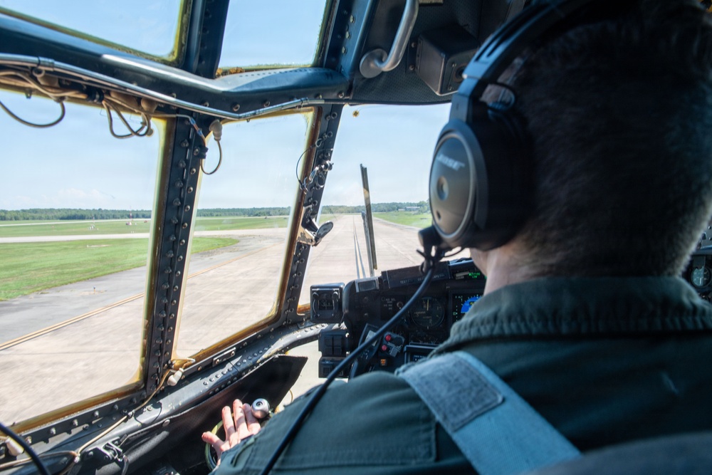 A pilot is seated in the cockpit of a large military aircraft, viewed from behind, taxiing or preparing for takeoff on a runway. The cockpit features aged, riveted window frames with a yellowed tint, suggesting an older aircraft design. The pilot is wearing Bose aviation headphones and a green flight suit, with instrument panels visible to the right.