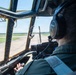 A pilot is seated in the cockpit of a large military aircraft, viewed from behind, taxiing or preparing for takeoff on a runway. The cockpit features aged, riveted window frames with a yellowed tint, suggesting an older aircraft design. The pilot is wearing Bose aviation headphones and a green flight suit, with instrument panels visible to the right.