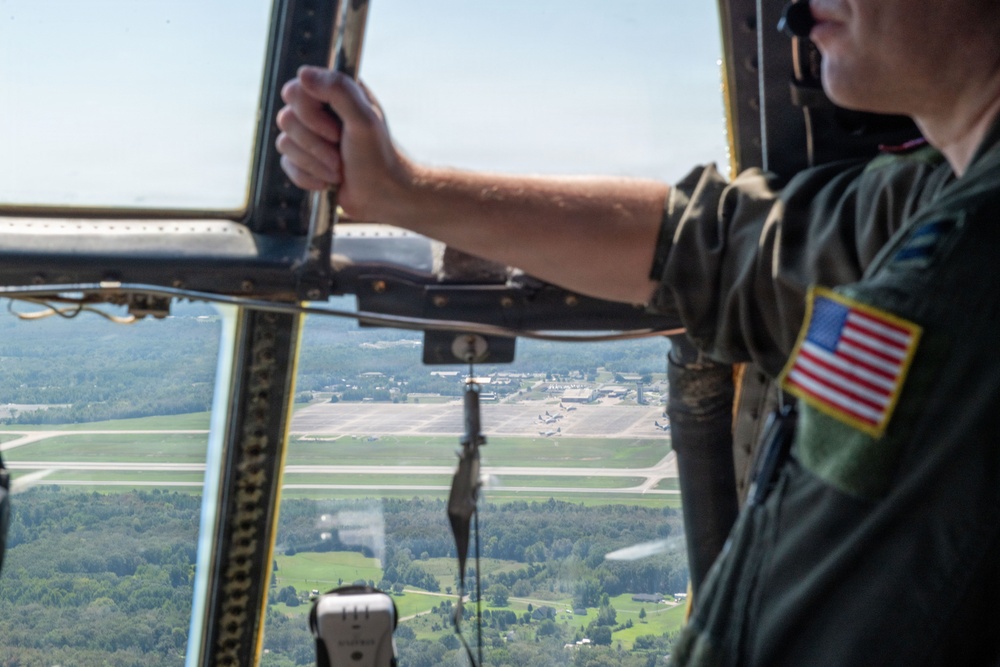 A U.S. military aircrew member in an olive green flight suit is looking out of an open aircraft window or door at altitude, observing an airfield below. The person wears a U.S. flag patch on their right shoulder, indicating active duty or reserve military status. The aerial view shows runways, taxiways, parked aircraft, and surrounding forested terrain.