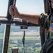 A U.S. military aircrew member in an olive green flight suit is looking out of an open aircraft window or door at altitude, observing an airfield below. The person wears a U.S. flag patch on their right shoulder, indicating active duty or reserve military status. The aerial view shows runways, taxiways, parked aircraft, and surrounding forested terrain.