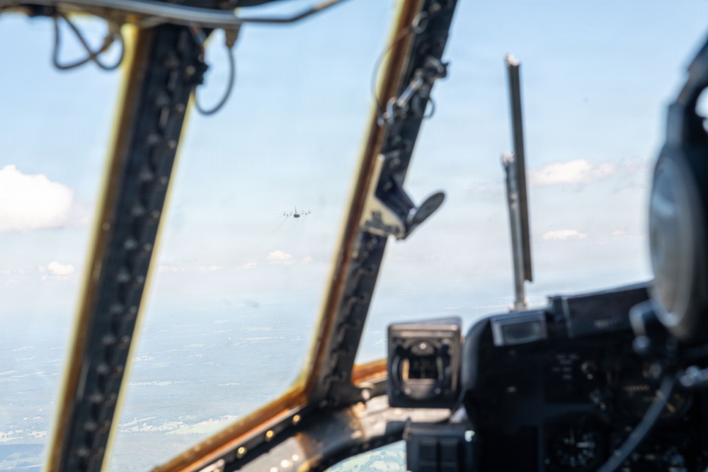 The image is taken from inside a military aircraft cockpit, looking forward through the windshield framing. A second aircraft is visible in the distance flying ahead, with terrain and scattered clouds visible below. The cockpit interior shows structural framing, instrument panels, and equipment mounts in the foreground.