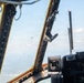 The image is taken from inside a military aircraft cockpit, looking forward through the windshield framing. A second aircraft is visible in the distance flying ahead, with terrain and scattered clouds visible below. The cockpit interior shows structural framing, instrument panels, and equipment mounts in the foreground.