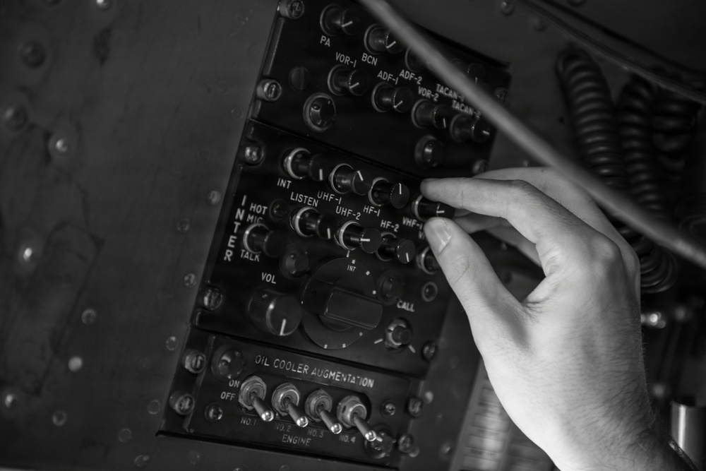 A black-and-white photograph shows a human hand operating controls on a vintage military aircraft communications and avionics panel. The panel contains multiple labeled rotary knobs, toggle switches, and selector controls for various radio and navigation systems. A lower panel is labeled 'Oil Cooler Augmentation' with numbered engine toggle switches.