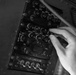 A black-and-white photograph shows a human hand operating controls on a vintage military aircraft communications and avionics panel. The panel contains multiple labeled rotary knobs, toggle switches, and selector controls for various radio and navigation systems. A lower panel is labeled 'Oil Cooler Augmentation' with numbered engine toggle switches.