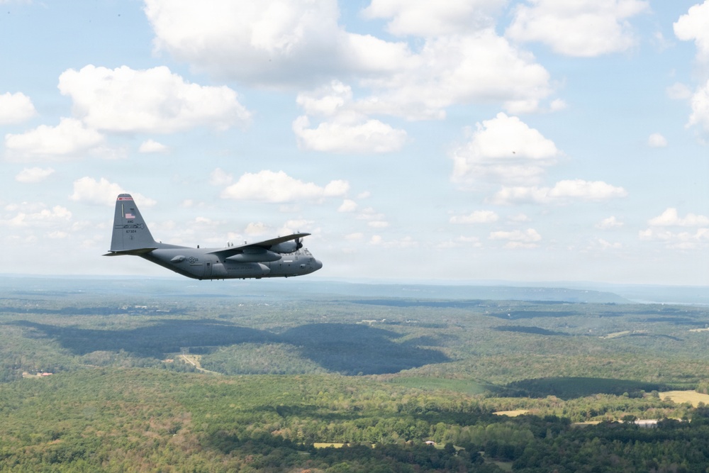 A gray military transport aircraft, consistent with a C-130 Hercules, is photographed in flight over a densely forested, hilly landscape under a partly cloudy sky. The aircraft bears a U.S. flag marking on its tail fin and appears to be cruising at moderate altitude. The photo was likely taken from another aircraft flying in formation or proximity.