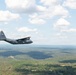 A gray military transport aircraft, consistent with a C-130 Hercules, is photographed in flight over a densely forested, hilly landscape under a partly cloudy sky. The aircraft bears a U.S. flag marking on its tail fin and appears to be cruising at moderate altitude. The photo was likely taken from another aircraft flying in formation or proximity.