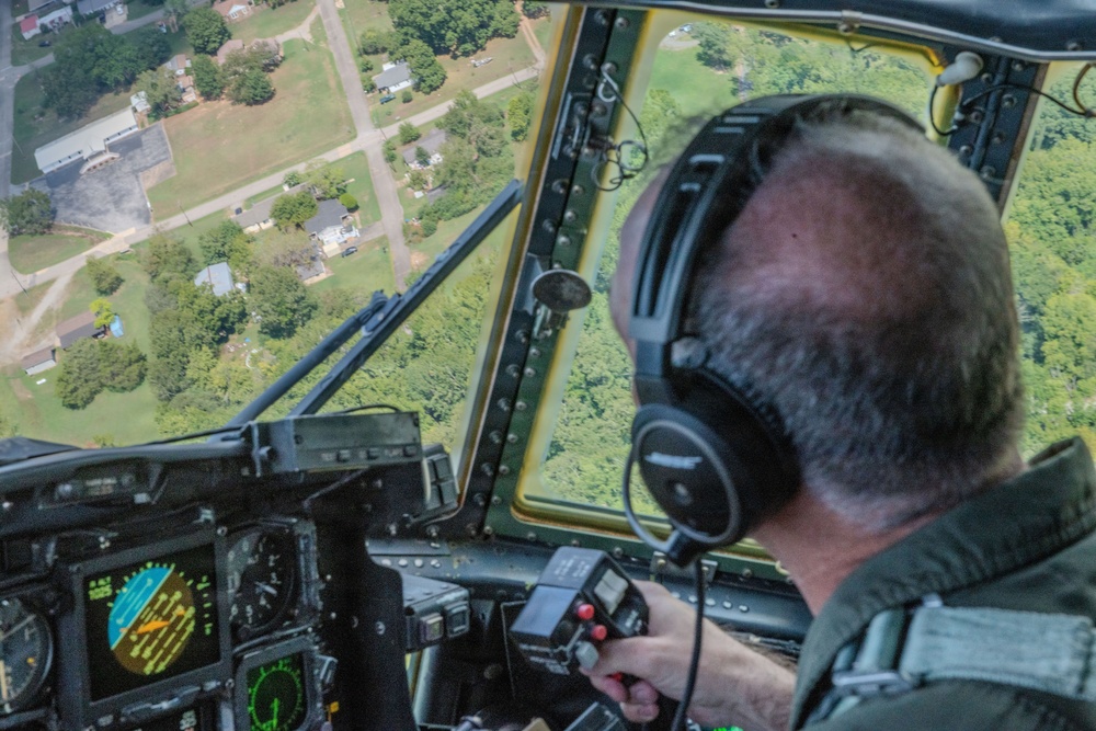 A military or aviation pilot is seated in the cockpit of a large aircraft, viewed from behind, flying over a suburban or semi-rural area. The pilot is wearing a flight suit and Bose aviation headset while holding a control stick with red buttons. The cockpit features multiple instrument displays and the view through the windshield shows residential neighborhoods and tree-covered terrain below.