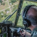 A military or aviation pilot is seated in the cockpit of a large aircraft, viewed from behind, flying over a suburban or semi-rural area. The pilot is wearing a flight suit and Bose aviation headset while holding a control stick with red buttons. The cockpit features multiple instrument displays and the view through the windshield shows residential neighborhoods and tree-covered terrain below.