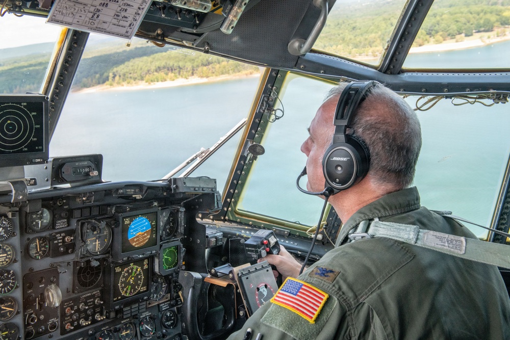 A U.S. military pilot is seated in the cockpit of a large military aircraft, likely a C-130 Hercules, flying over a body of water surrounded by forested terrain. The pilot is wearing an olive drab flight suit with a U.S. flag patch and Bose aviation headset while operating flight controls. The cockpit displays a mix of analog gauges and modern digital avionics screens.