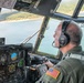 A U.S. military pilot is seated in the cockpit of a large military aircraft, likely a C-130 Hercules, flying over a body of water surrounded by forested terrain. The pilot is wearing an olive drab flight suit with a U.S. flag patch and Bose aviation headset while operating flight controls. The cockpit displays a mix of analog gauges and modern digital avionics screens.