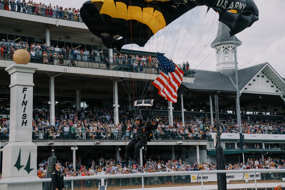 A U.S. Army parachutist is landing at Churchill Downs racetrack, carrying an American flag during what appears to be a pre-race ceremony. The parachutist is descending near the iconic 'FINISH' line post with black and gold U.S. Army branded parachute canopies visible overhead. The grandstands are packed with thousands of spectators, consistent with a major event such as the Kentucky Derby.