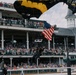 A U.S. Army parachutist is landing at Churchill Downs racetrack, carrying an American flag during what appears to be a pre-race ceremony. The parachutist is descending near the iconic 'FINISH' line post with black and gold U.S. Army branded parachute canopies visible overhead. The grandstands are packed with thousands of spectators, consistent with a major event such as the Kentucky Derby.