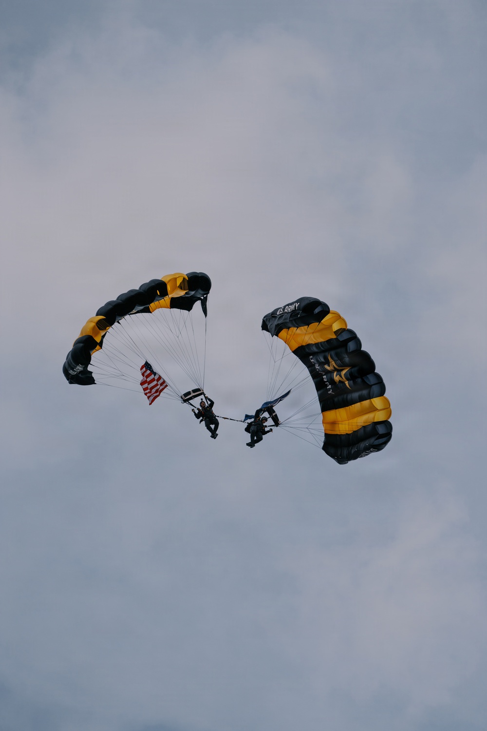 Two military skydivers are descending under black and gold parachutes marked with 'U.S. ARMY' branding, consistent with the U.S. Army Golden Knights parachute team. One jumper is carrying an American flag during the descent. The image is taken from a low angle against an overcast sky.